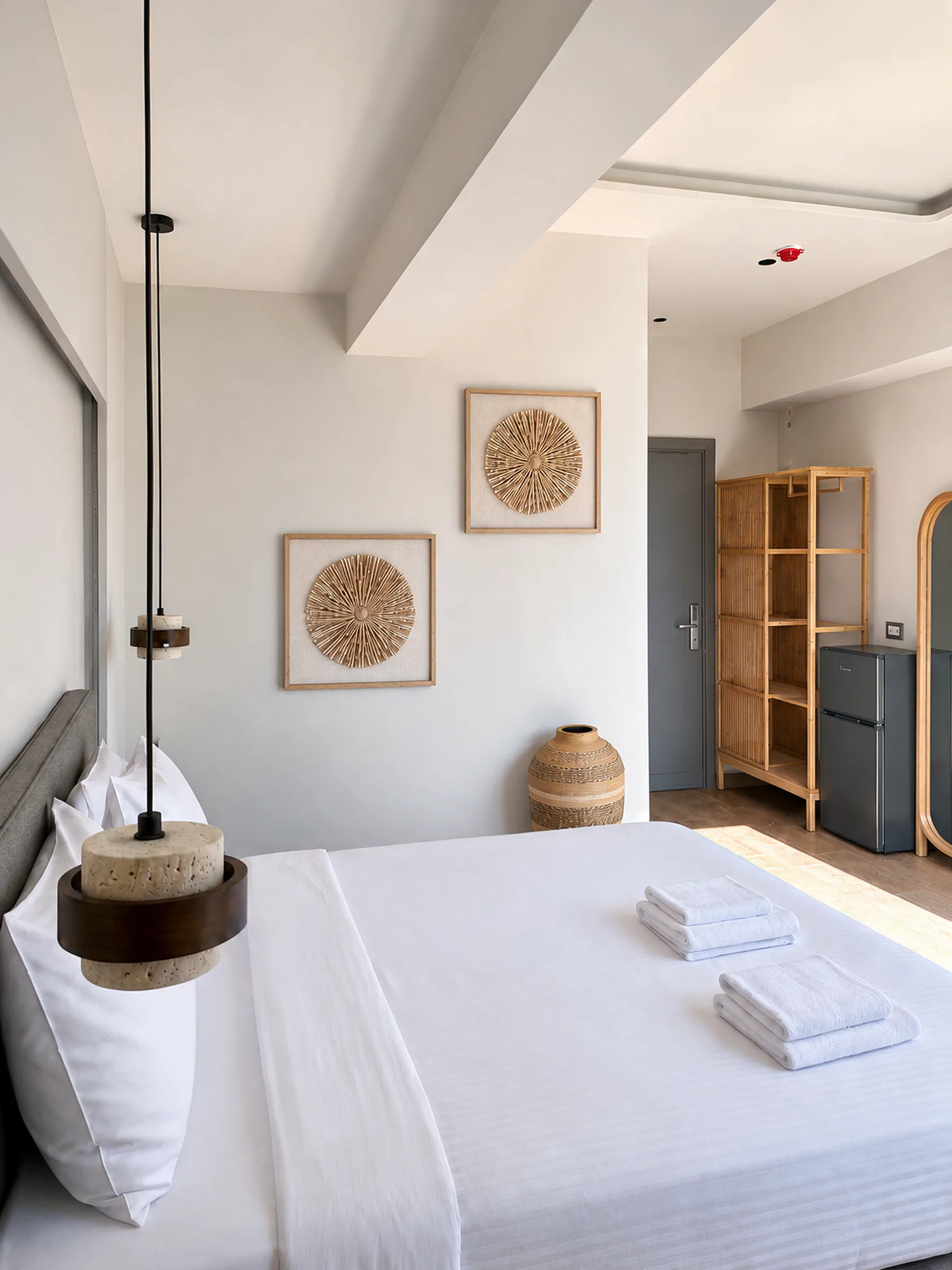 A double bed with white linen and folded towels, two framed woven artworks above and a pendant lamp at the bedside