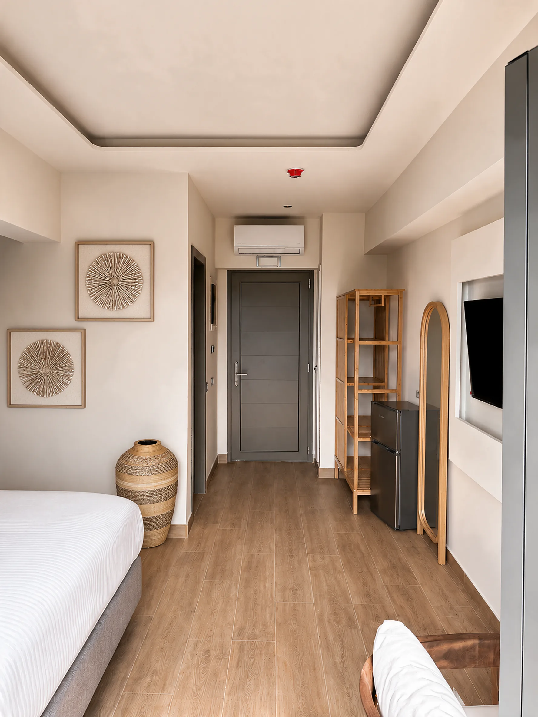 The room looking towards the door, with open wooden shelving, a mini fridge, an arched full-length mirror and a woven floor vase