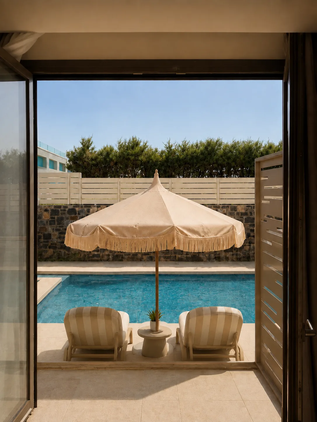 Looking out of a room to the pool, a cream fringed parasol and two loungers against the stone wall and hedge